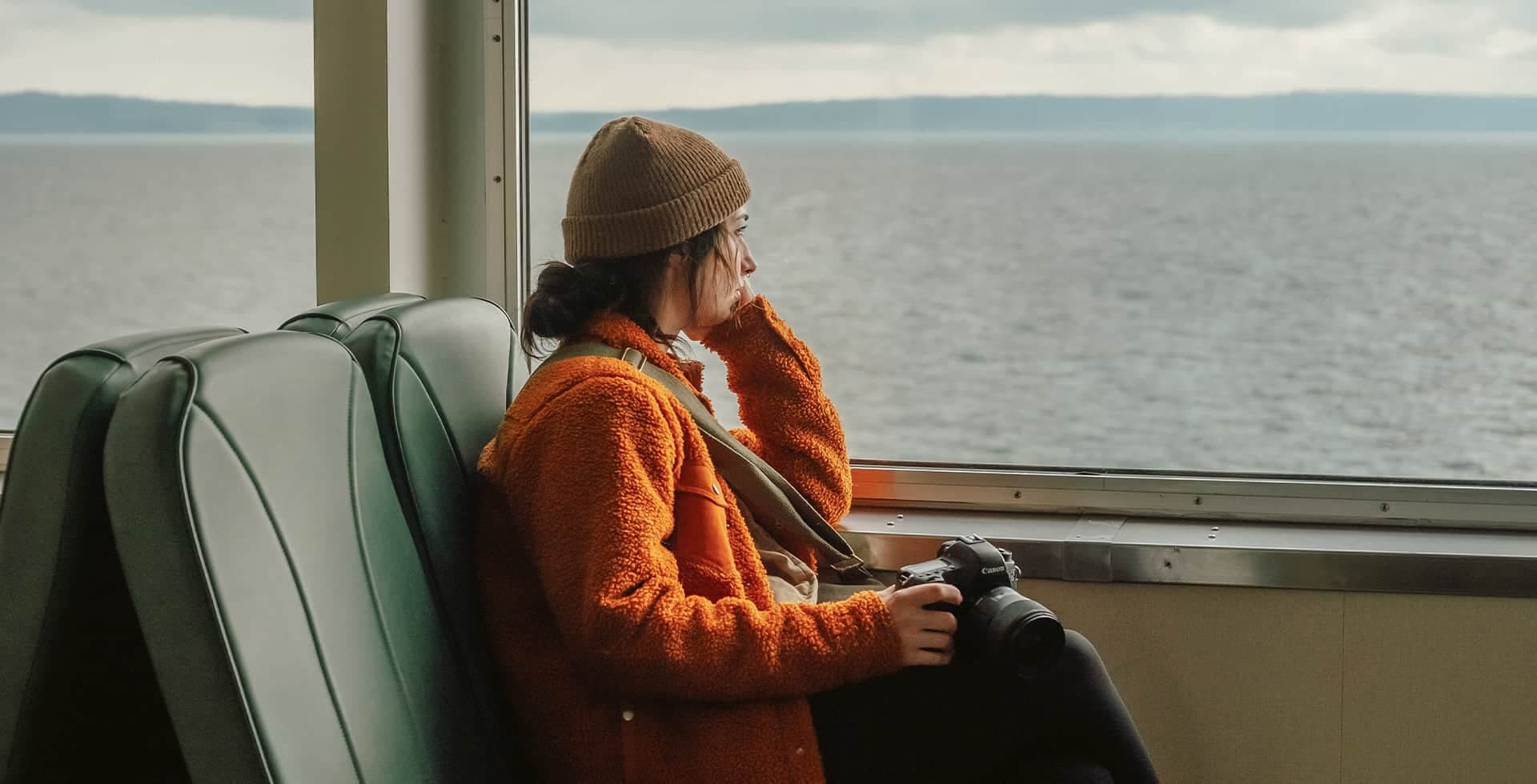 En ung kvinna sitter ensam och stirrar ut genom ett fönster mot vattnet.